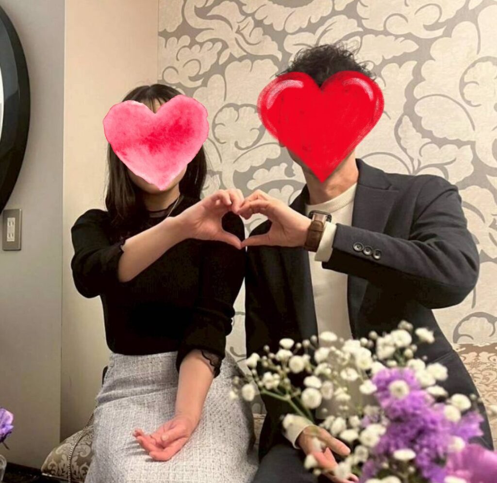 花飾りの前でふたりのそれぞれの手でハートマークをつくるカップル